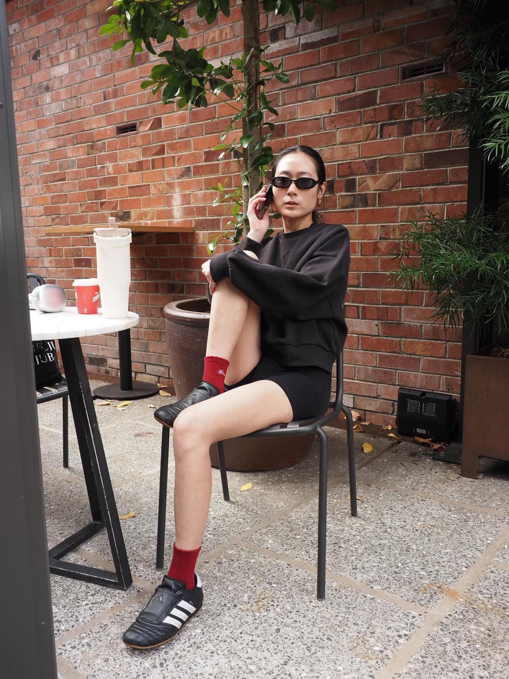 A woman wearing a black long-sleeve top, black shorts, black Adidas soccer cleats, and red socks sits on a black metal chair in front of a red brick wall.