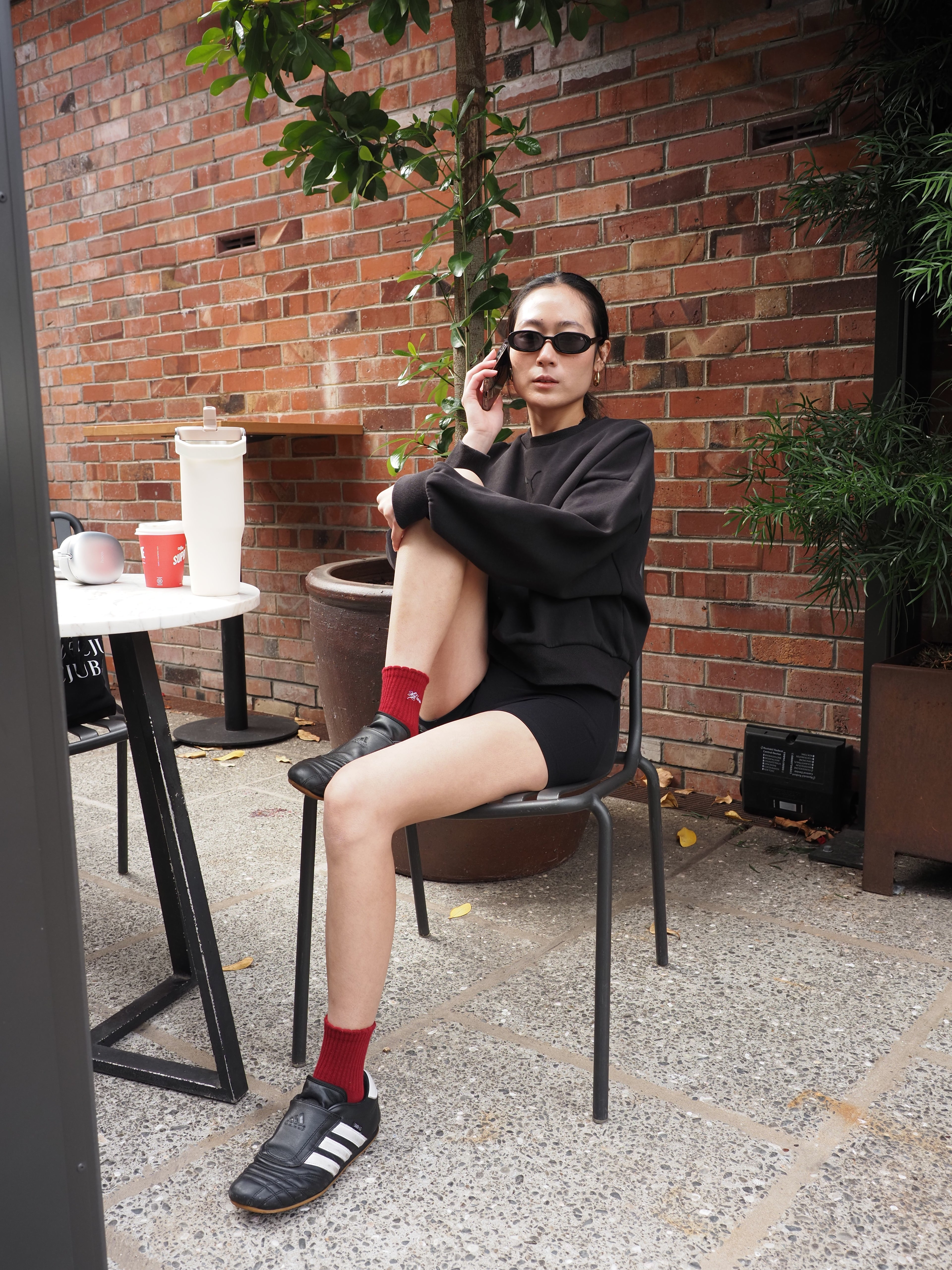 A woman wearing a black long-sleeve top, black shorts, black Adidas soccer cleats, and red socks sits on a black metal chair in front of a red brick wall.
