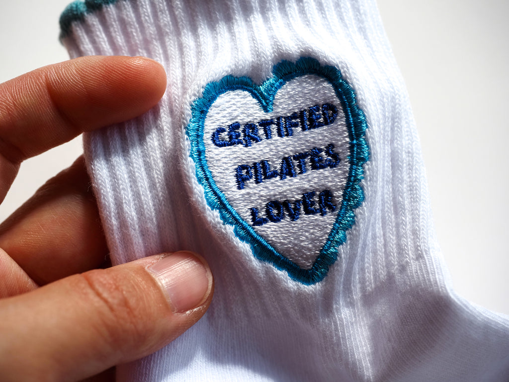 A close-up of a white grip sock, featuring a heart-shaped embroidered patch with a blue border and the text 'CERTIFIED PILATES LOVER'.