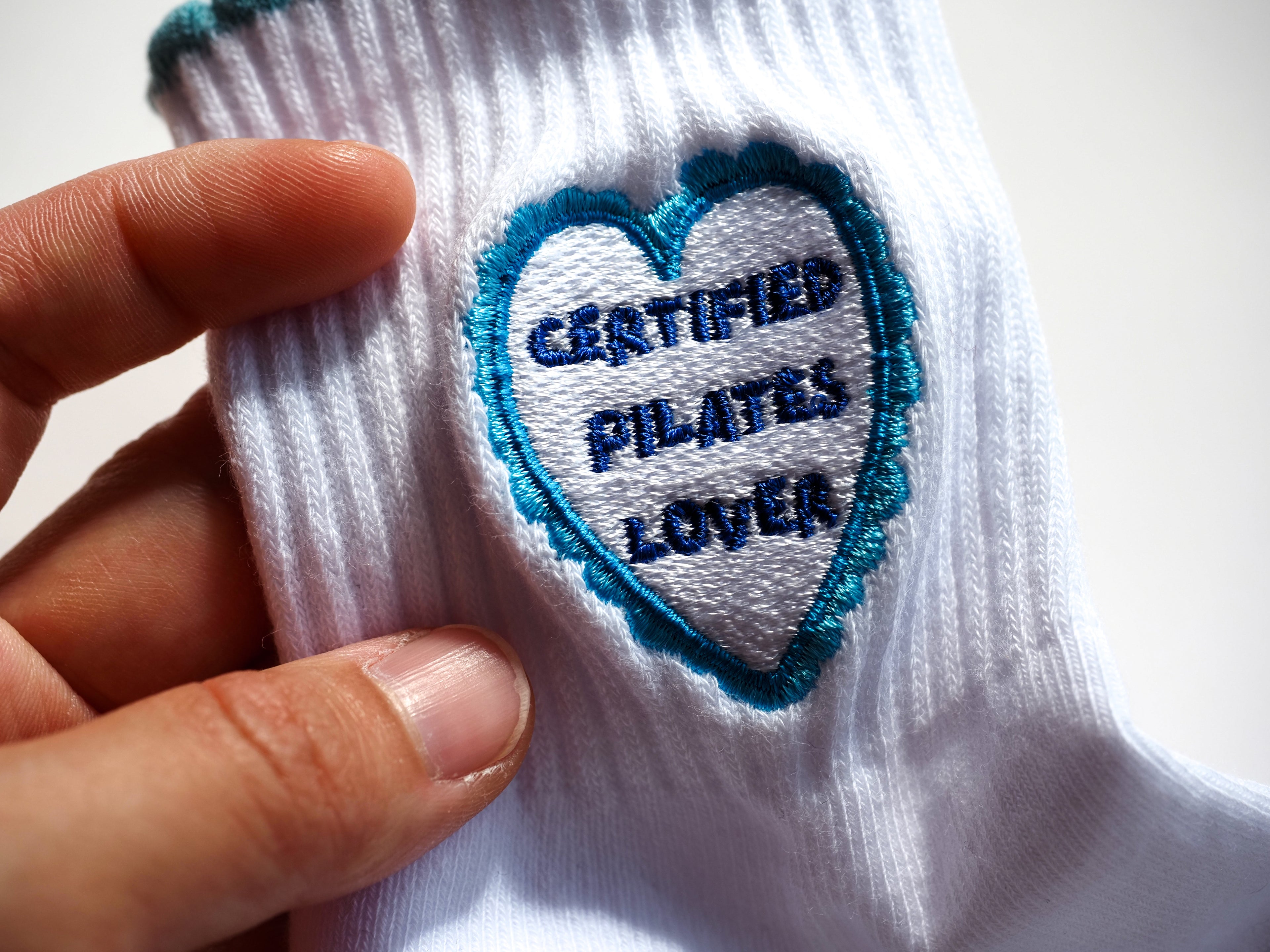 A close-up of a white grip sock, featuring a heart-shaped embroidered patch with a blue border and the text 'CERTIFIED PILATES LOVER'.