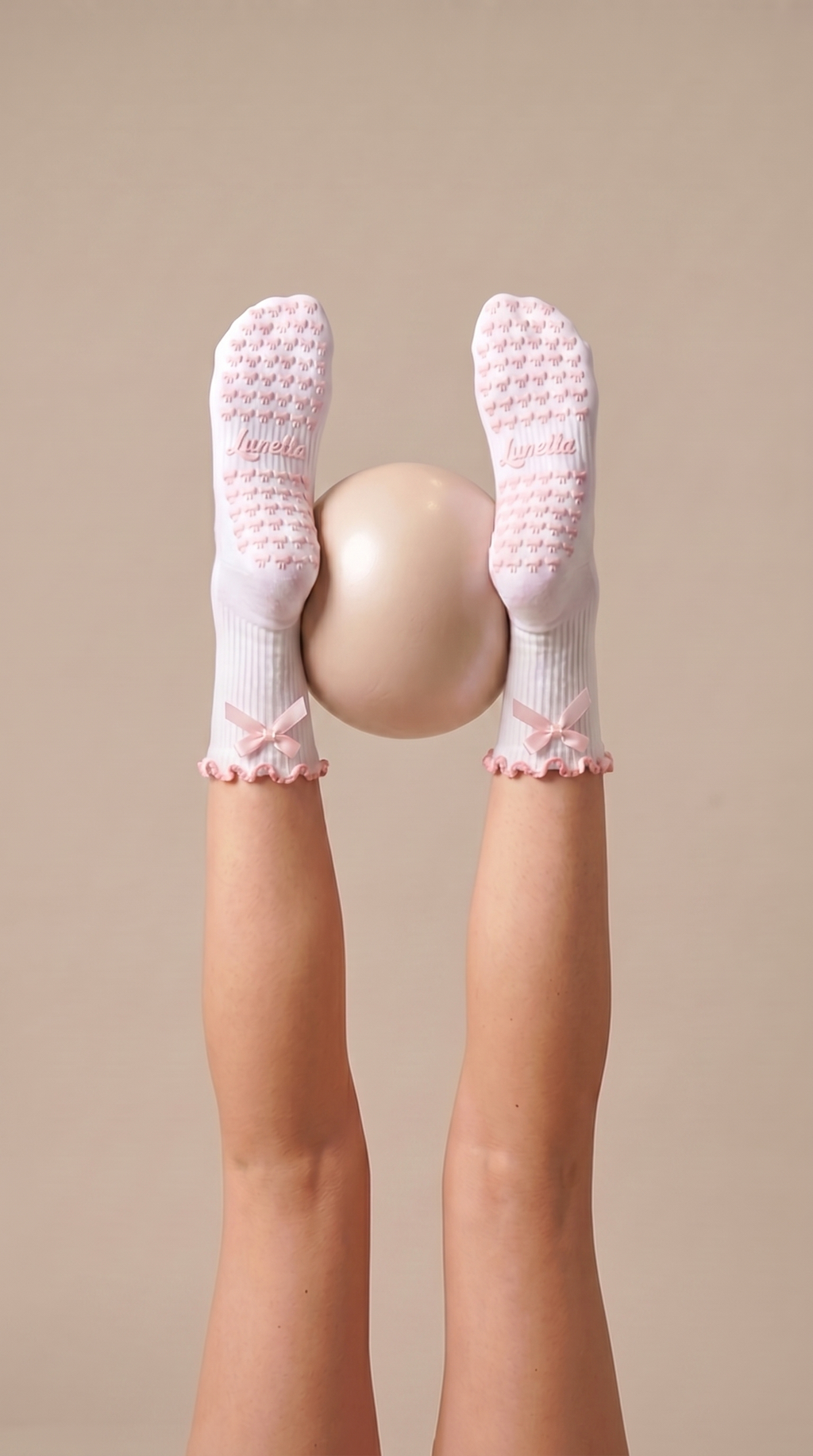 Aesthetic, white pilates grip socks with pink ribbon on feet holding a beige exercise ball in reformer class