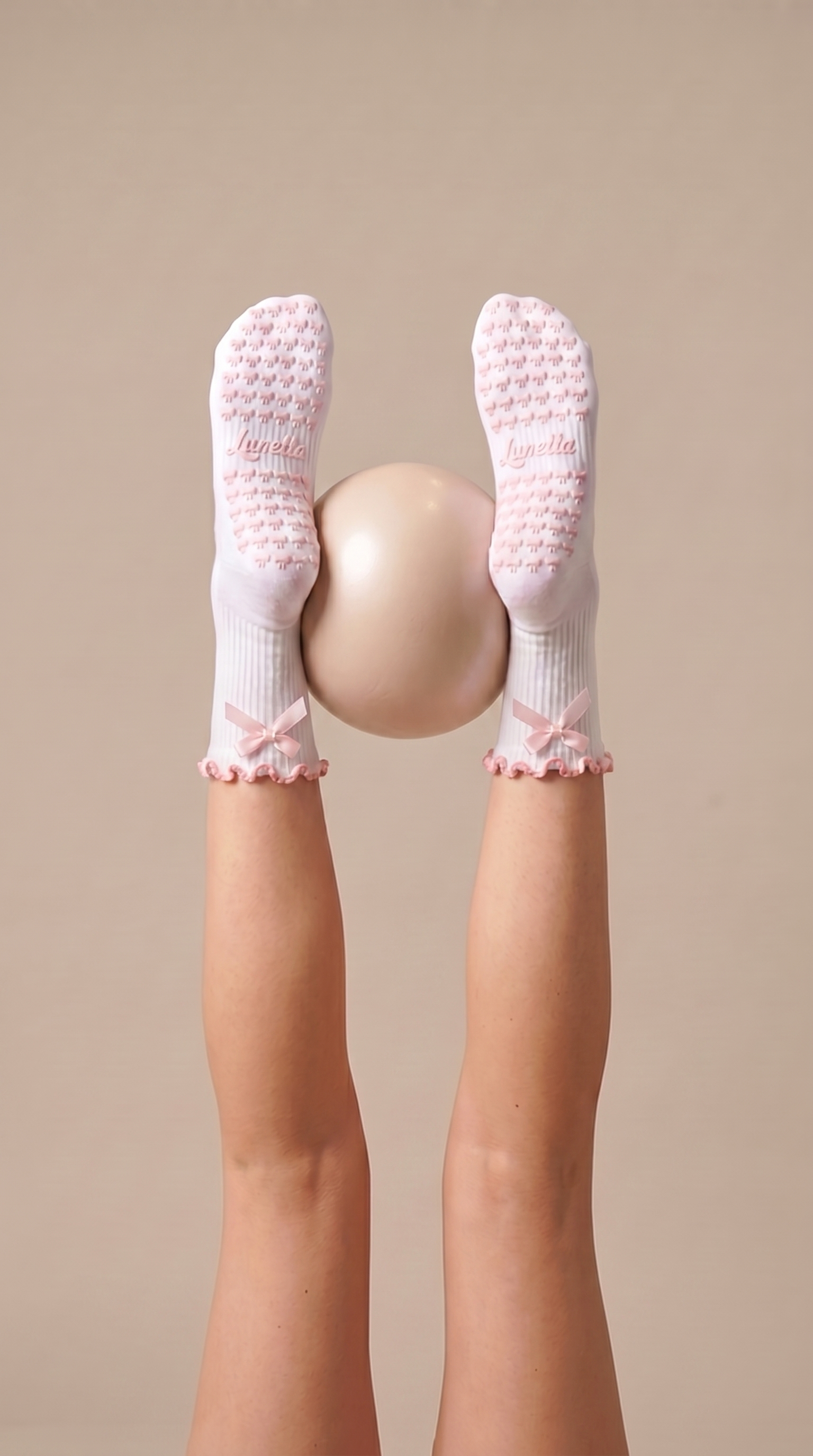 Aesthetic, white pilates grip socks with pink ribbon on feet holding a beige exercise ball in reformer class