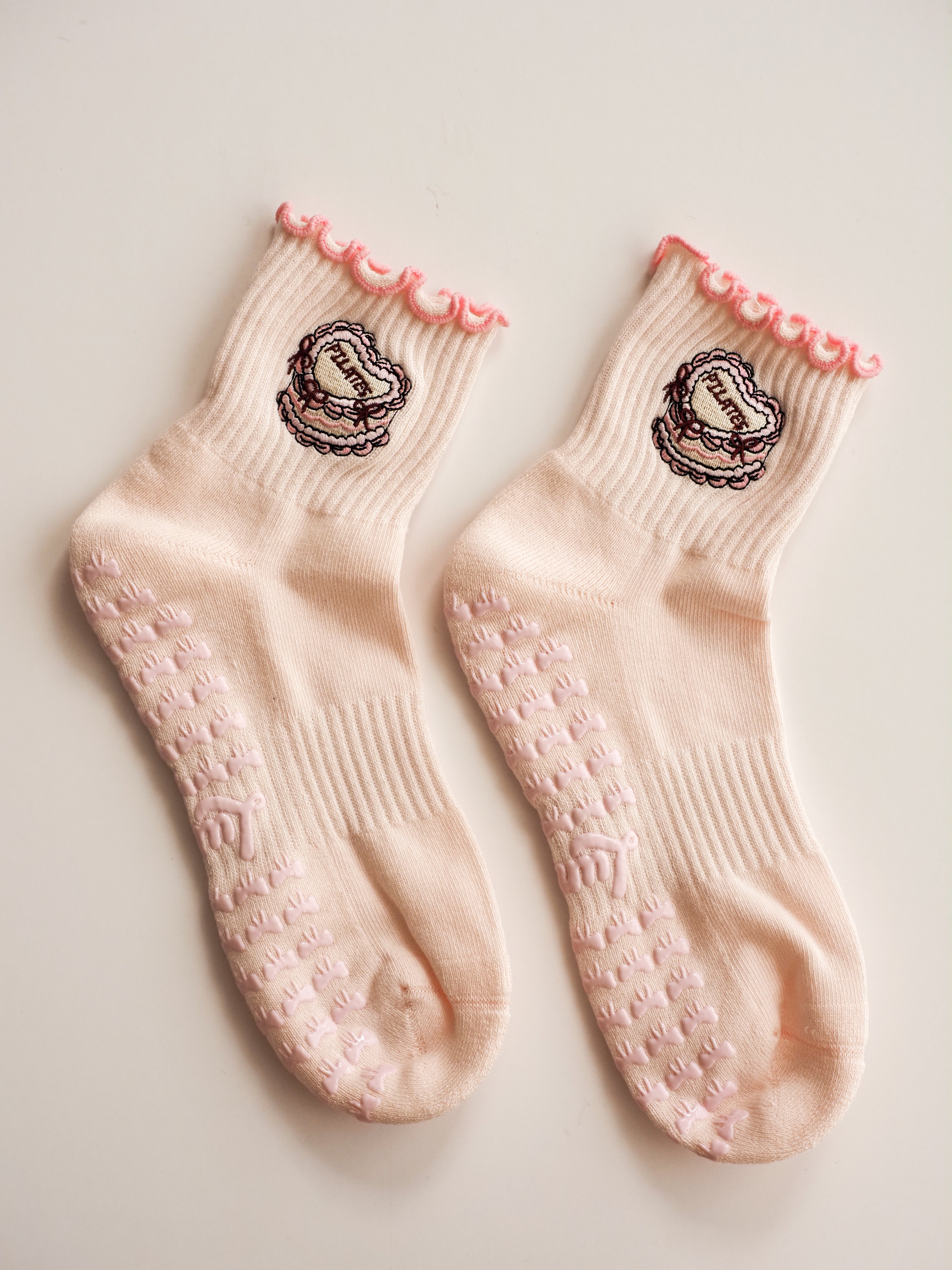 Product shot of a grip sock in pale blush pink, featuring a scalloped pink trim at the top and a central embroidered cake.