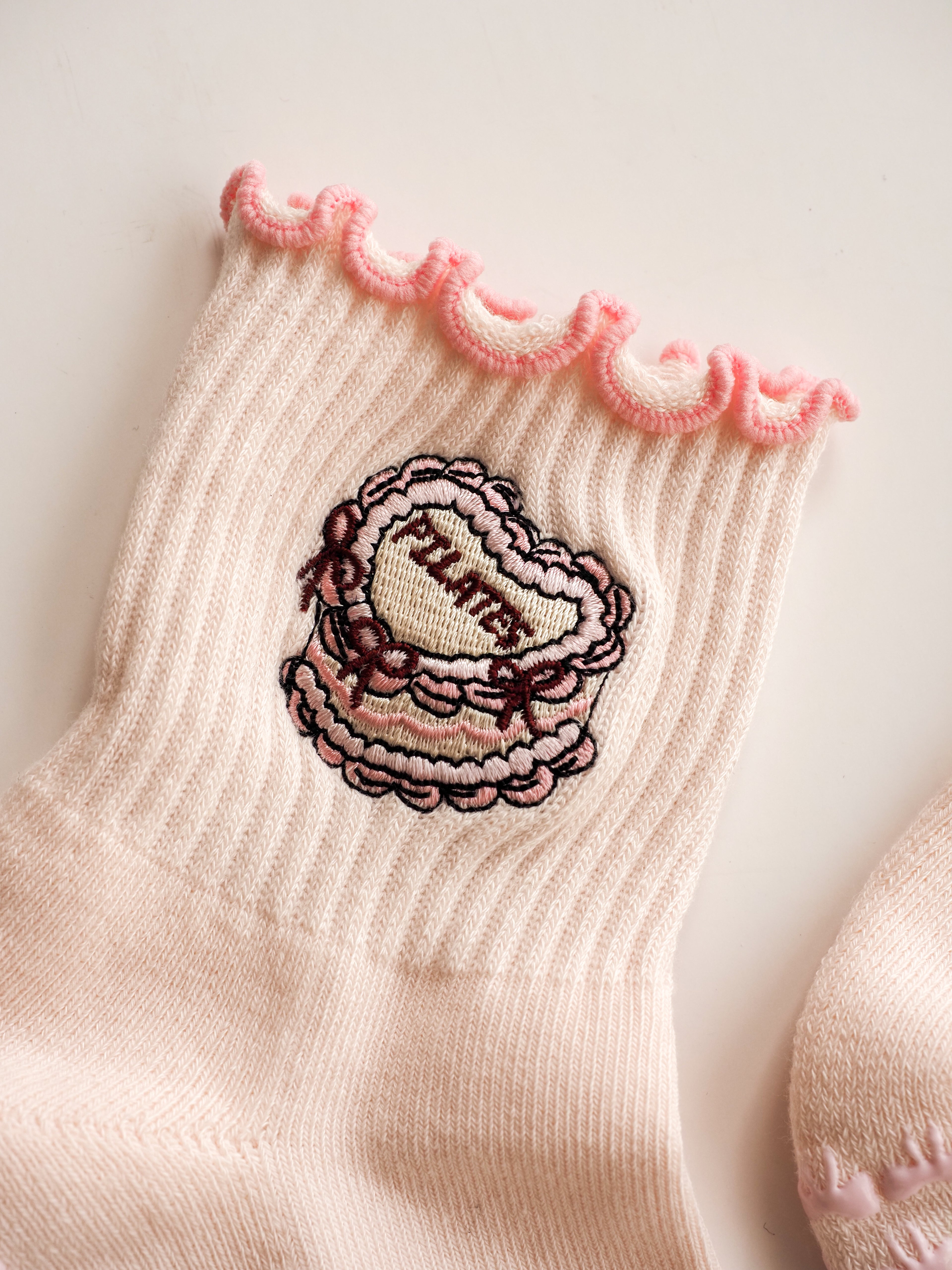 Close-up of a grip sock in pale blush pink, featuring a scalloped pink trim at the top and a central embroidered cake.